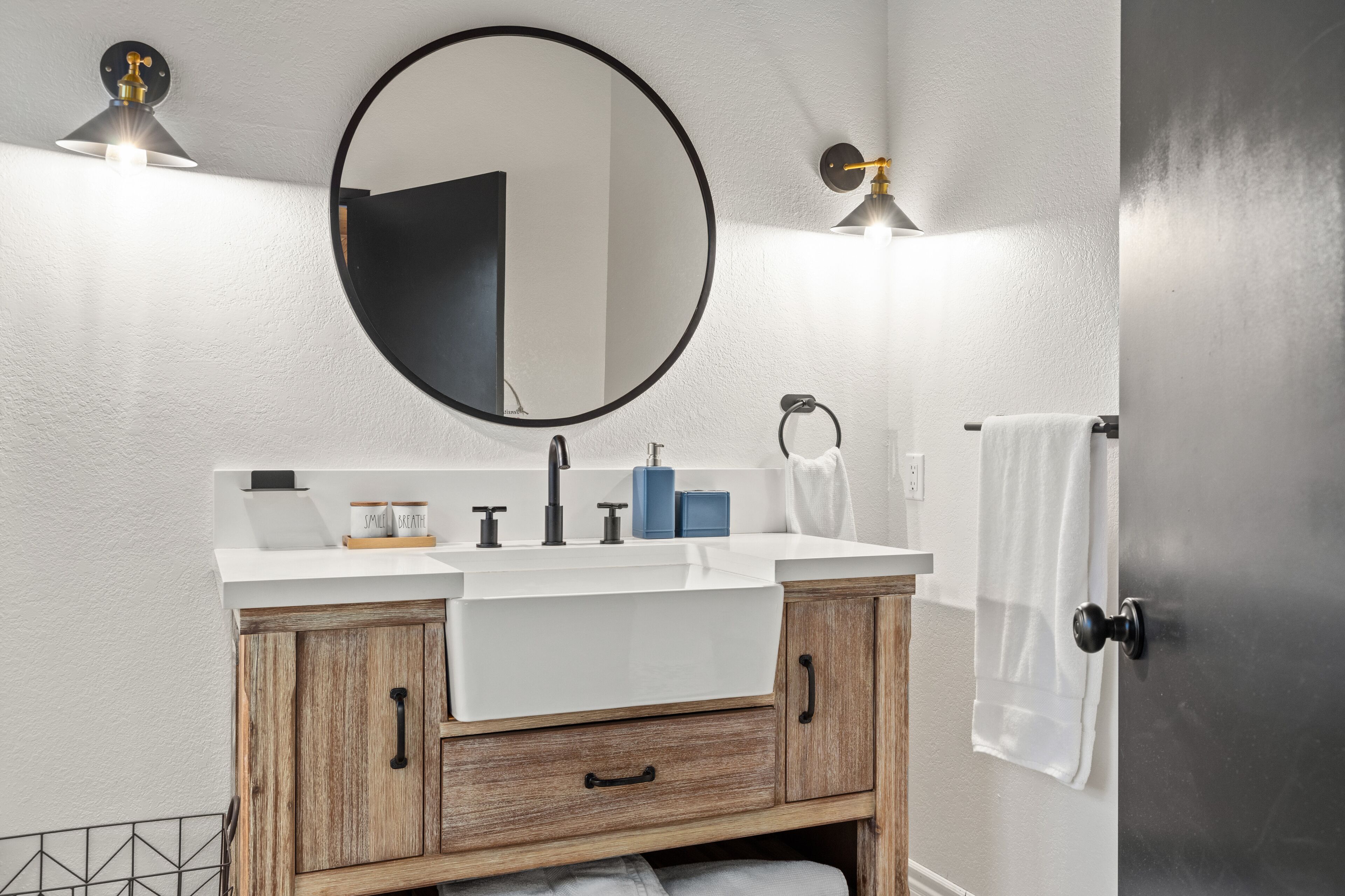 Een badkamer met een wit wastafelmeubel, houten kastjes, een ronde spiegel en twee wandlampen.