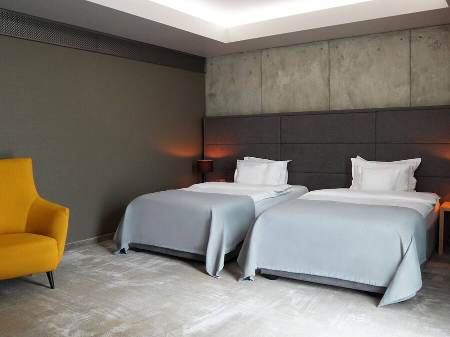 Habitación de hotel con dos camas, un sillón amarillo y una pared de concreto como detalle.