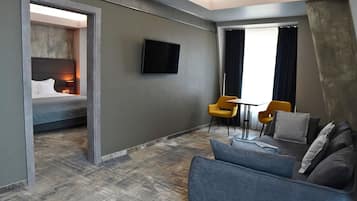 A modern hotel room with a bed, a sofa, two yellow chairs, a TV, and a large window with curtains.