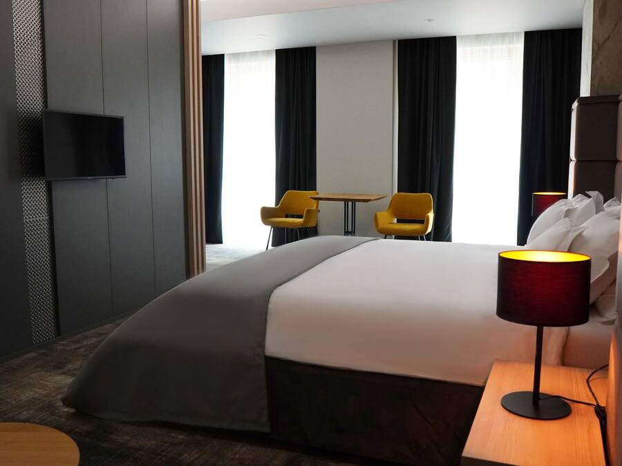 Una habitación de hotel moderna con una cama grande, un televisor montado en la pared, un escritorio con una lámpara y dos sillas.