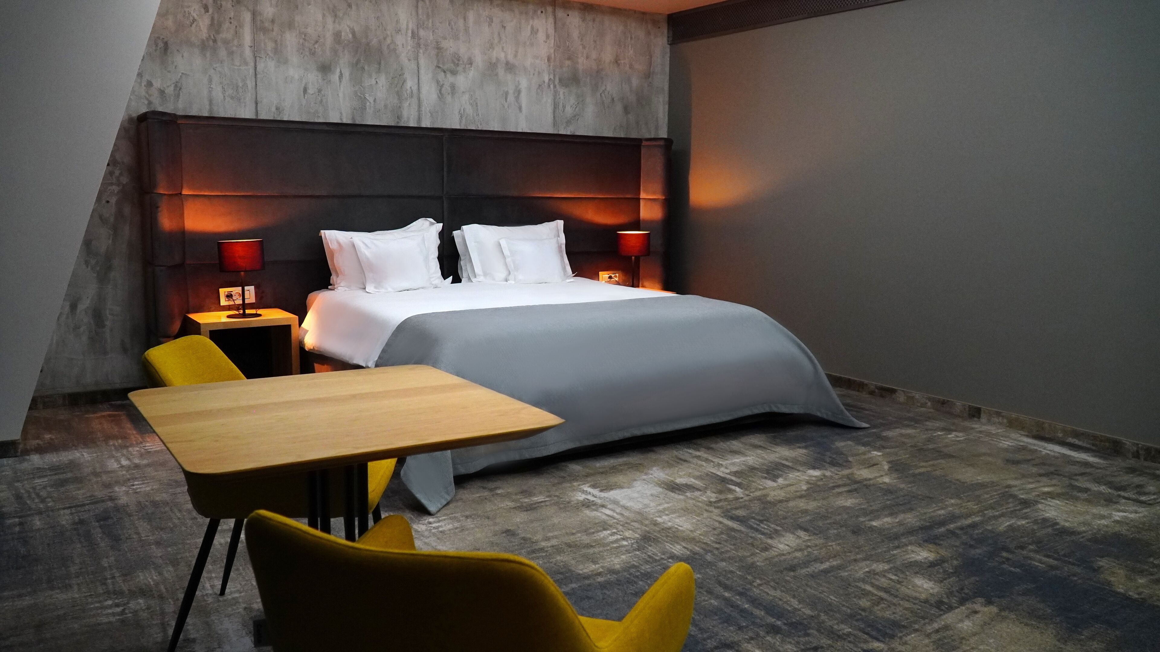 Una habitación de hotel moderna con una cama grande, dos mesitas de noche, un escritorio de madera y una silla amarilla.