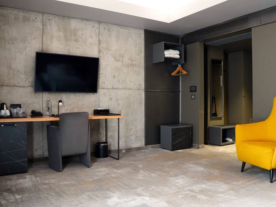 A modern hotel room with a large flat-screen TV mounted on a concrete wall, a desk with a chair, a small cabinet, and a bright yellow armchair.