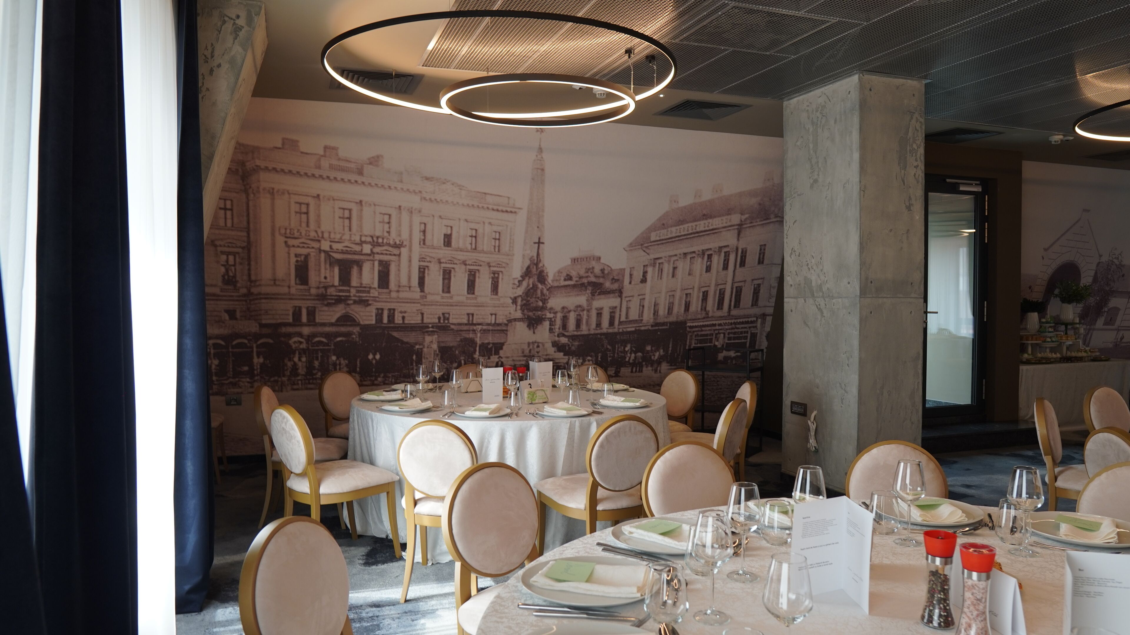 Een restaurant met ronde tafels gedekt voor diners, een muurschildering van een stadsgezicht en moderne verlichtingsarmaturen.