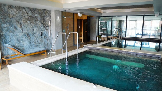 An indoor swimming pool with a wooden bench, a glass wall, and a view of the outside. - Aniroc Signature Hotel (Arad)
