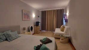 A hotel room with a bed, a sofa, a small table, and a curtain. - Spacious Studio Apartment overlooking garden and pool area (Albufeira)
