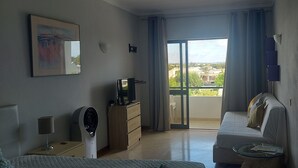 A hotel room with a bed, a sofa, a TV on a dresser, a fan, a window with curtains, and a view of the outside. - Spacious Studio Apartment overlooking garden and pool area (Albufeira)