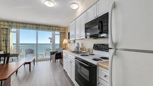 A modern kitchen with white cabinets, a black microwave, and a stove. There is a dining table with chairs and a view of the ocean through large windows. - HS-1109 Oceanfront 1-Bedroom Condo in Myrtle Beach - Sleeps 6 (Myrtle Beach)