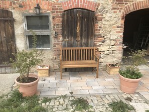 Un banc en bois se trouve devant un bâtiment rustique en briques, doté d’une grande porte en bois. De part et d’autre du banc, on peut voir des plantes en pot.