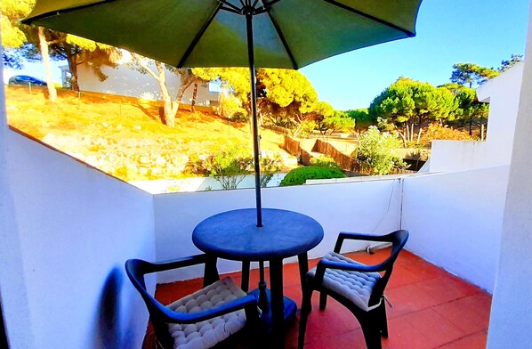 A balcony with a table and two chairs, an umbrella, and a view of a landscaped area with trees and a building in the background. - Villa Bella 200 meters from Falésia Beach by Urban Hypotenuse (Albufeira)