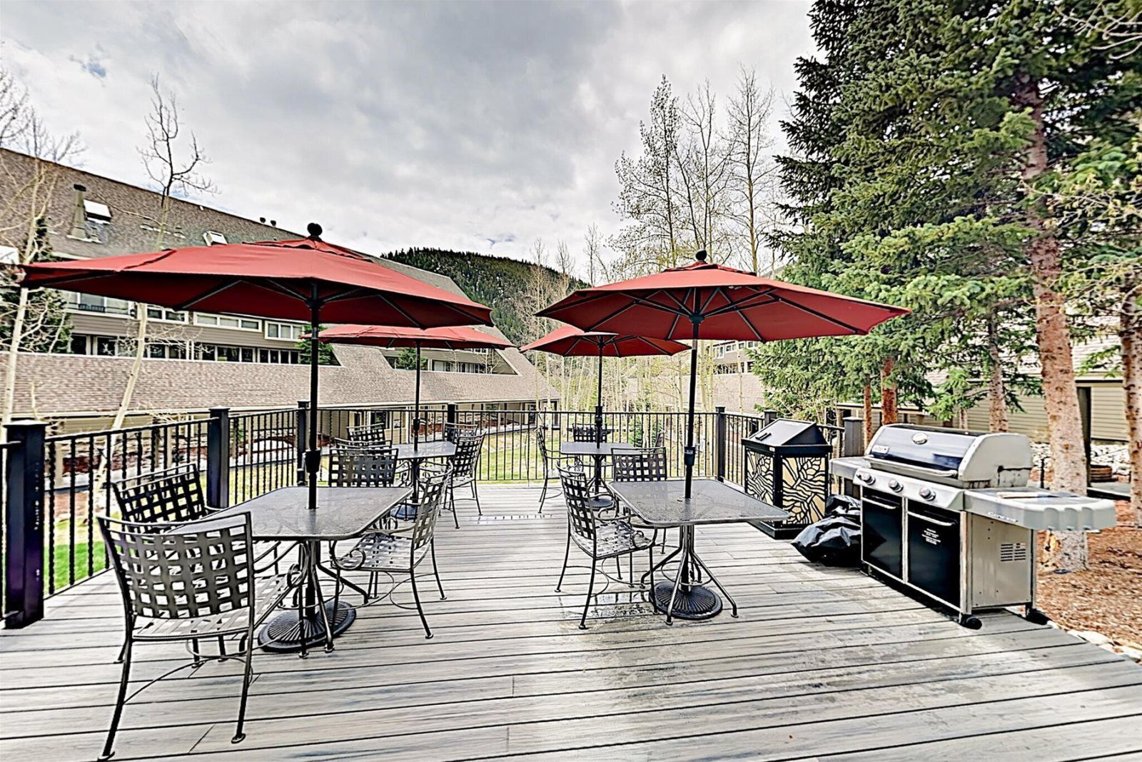 Un patio exterior con mesas y sillas, sombrillas rojas y una parrilla de barbacoa.