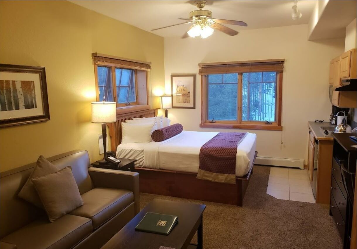 A hotel room with a bed, a sofa, a desk, a ceiling fan, and two windows.