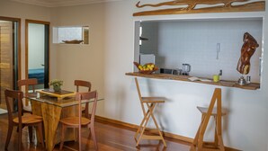 A dining area with a glass-top table, wooden chairs, and a wooden stool. A kitchen area is visible in the background with a hanging meat hook and a window above the sink. - APTO VISTA MAR MELHOR LOCALIZAÇÃO (Fortaleza)