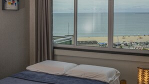A bedroom with a bed, a ceiling fan, a window with a view of the beach, and a lamp on a side table. - APTO VISTA MAR MELHOR LOCALIZAÇÃO (Fortaleza)