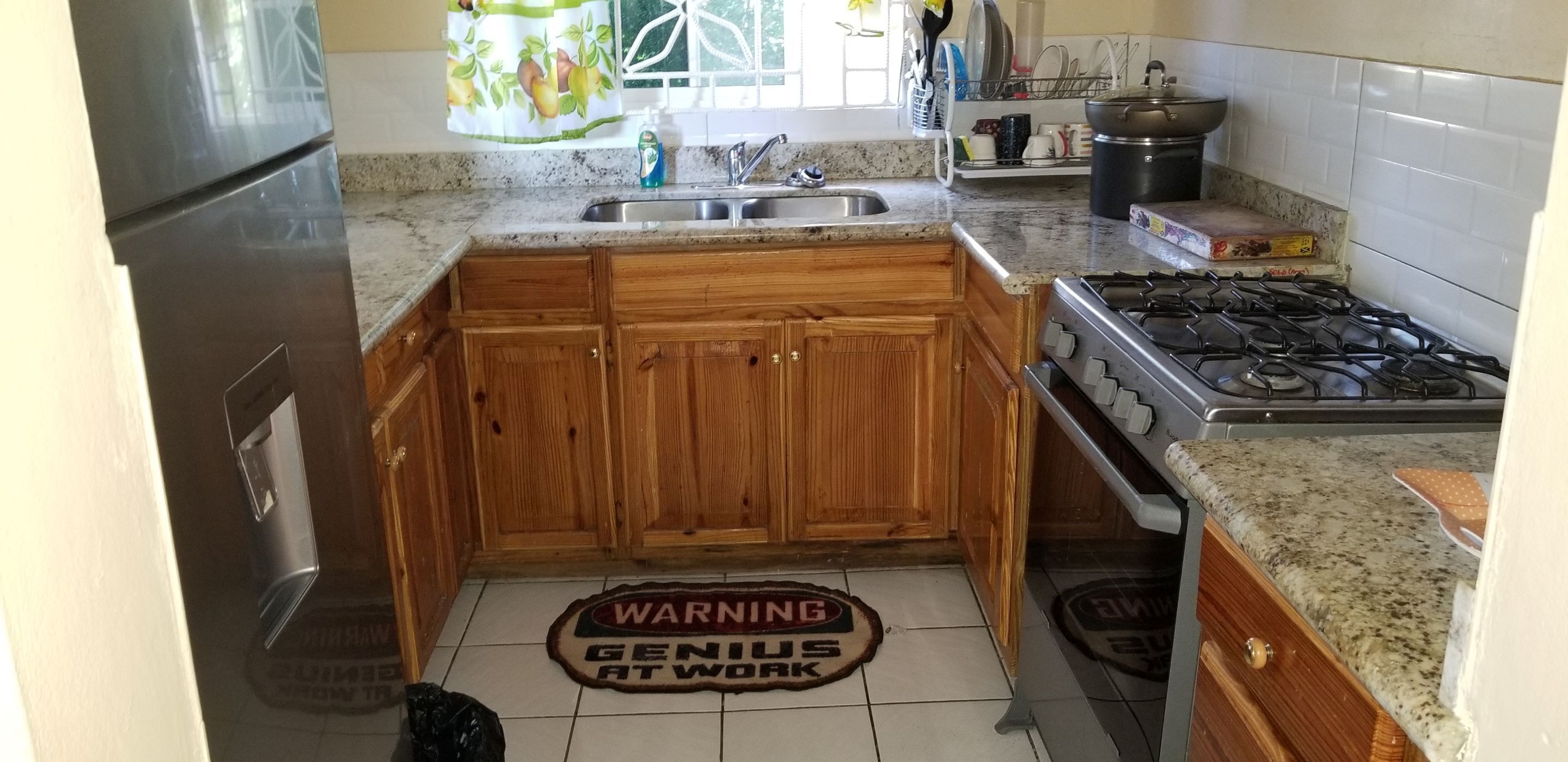 Una cocina con armarios de madera, un lavamanos de acero inoxidable, una estufa a gas y una ventana con cortina estampada de frutas.