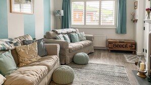 A cozy living room with two sofas, a floor lamp, a wooden chest labeled 'Beach', and a view of the outside through large windows. - 2 bedroom accommodation in Combe Martin (Combe Martin)