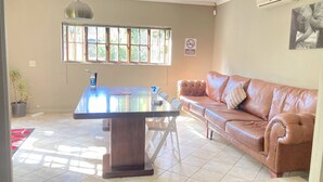 A living room with a brown leather sofa, a dining table with chairs, a window with blinds, and a 'No Smoking' sign on the wall.