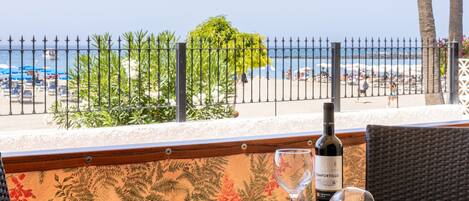 A table with a bottle of wine and a glass, set against a backdrop of a beach view, with a fence and foliage in the foreground.