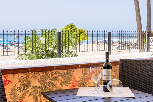 A table with a bottle of wine and a glass, set against a backdrop of a beach view, with a fence and foliage in the foreground.