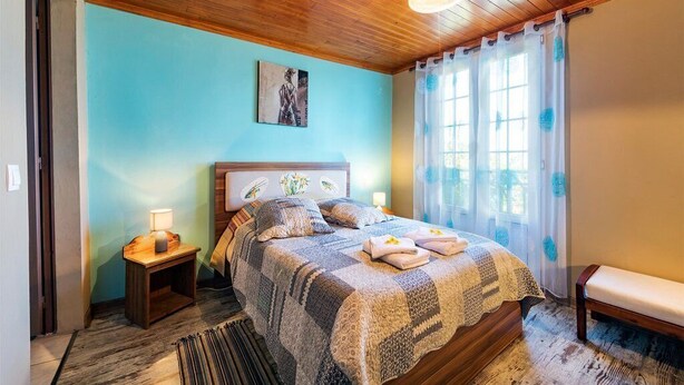 A bedroom with a wooden ceiling, a bed with a patterned blanket, a bedside table with a lamp, and a bench.