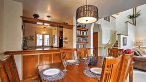 Zona pranzo con tavolo in legno apparecchiato per sei persone, circondato da sedie. È presente anche un angolo cottura con pensili in legno e un lampadario sopra il bancone.