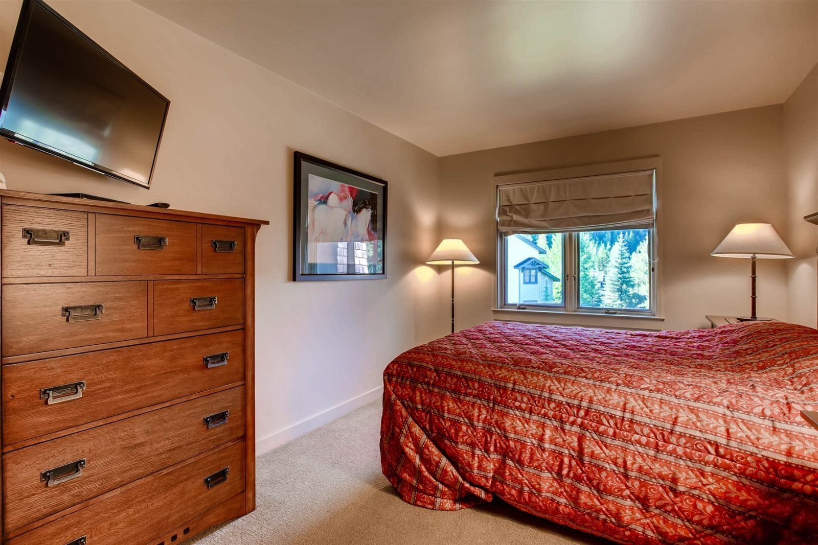 A bedroom with a large bed, a wooden dresser, a flat-screen TV, a window with a view, and two bedside lamps.
