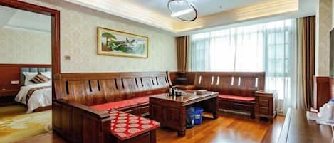 A hotel room with a wooden bench, a small table, a bed with a headboard, a large window with curtains, and a painting on the wall.