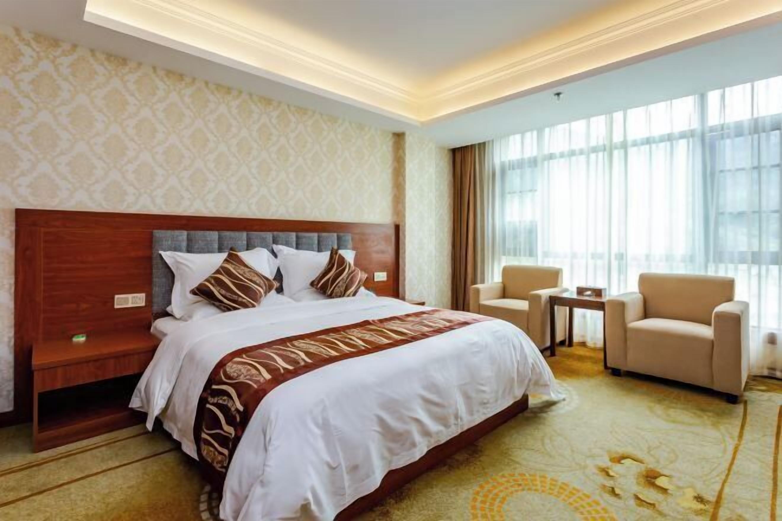 A hotel room with a large bed, a wooden headboard, a small table, two chairs, and a large window with curtains.