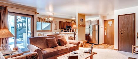A spacious living room with a sofa, coffee table, and chairs, a kitchen with modern appliances, and a large window offering a view of the outdoors.