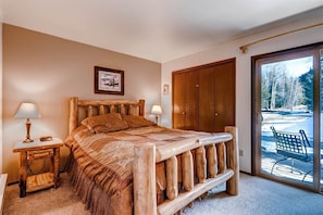 Une chambre avec un lit en bois, une table de chevet avec une lampe, un banc en bois à l’extérieur, et une vue sur un paysage enneigé par une porte vitrée.