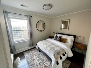 A bedroom with a bed, a nightstand, a mirror, a window with curtains, and a rug.