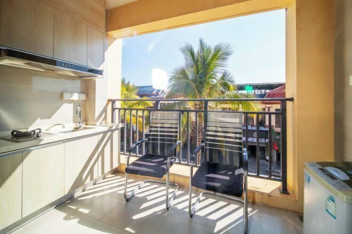 A kitchen with a window overlooking palm trees, a balcony with chairs, and a trash bin.