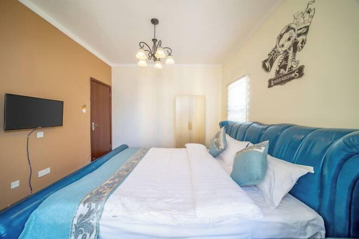 A bedroom with a bed, a blue upholstered headboard, a flat-screen TV, a chandelier, and a wall decoration.