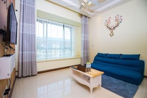A living room with a blue sofa, a small white table, a window with curtains, and a wall decoration. - Yijiayi Holiday Hotel (Jinghong)