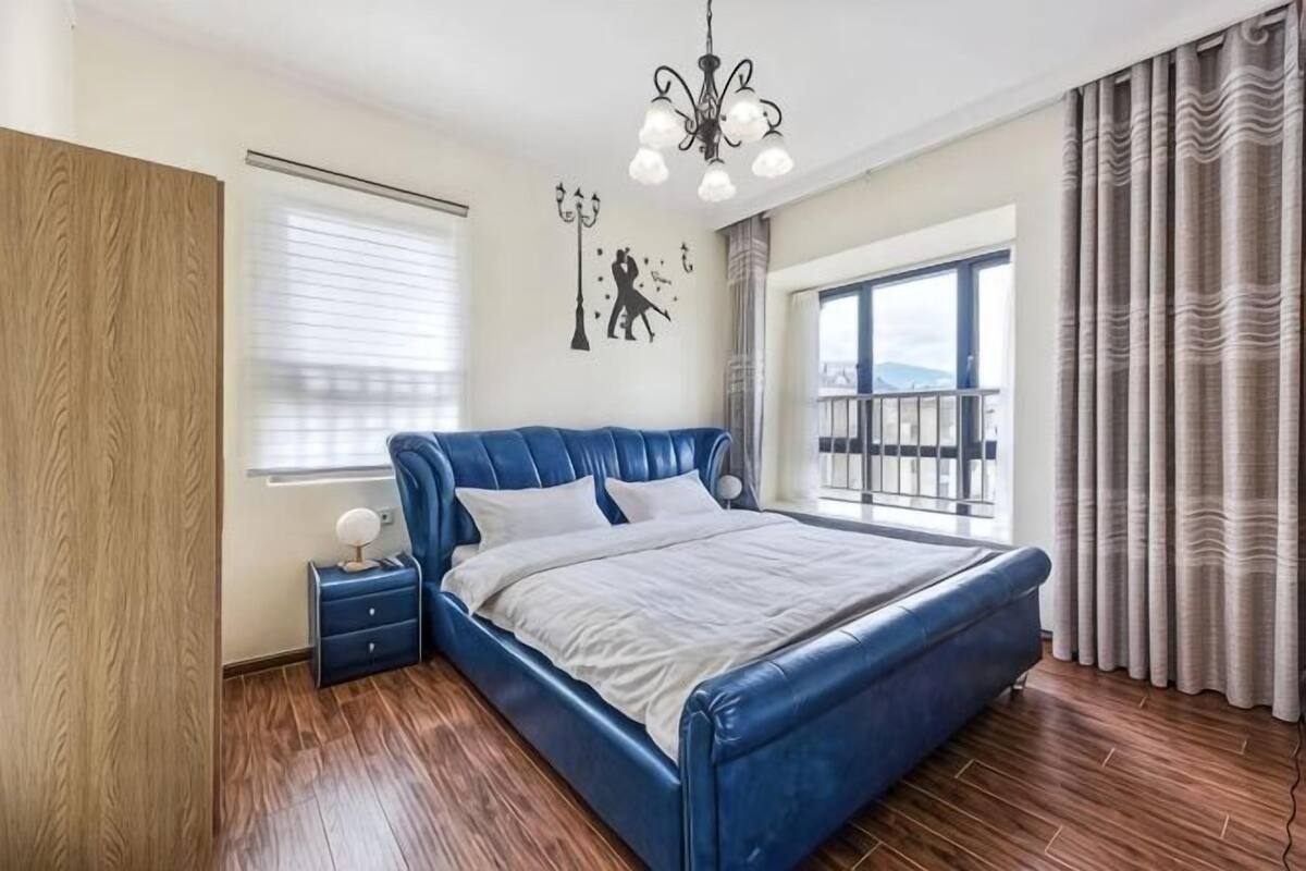 A bedroom with a large bed, a nightstand, a chandelier, a window with curtains, and a view of the outside.