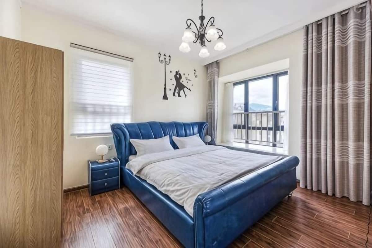 Une chambre à coucher avec un grand lit, une table de chevet, un lustre, une fenêtre avec des rideaux et une vue sur l’extérieur.