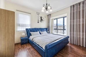 Un dormitorio con una cama grande, una mesita de noche, una araña, una ventana con cortinas y vistas al exterior.