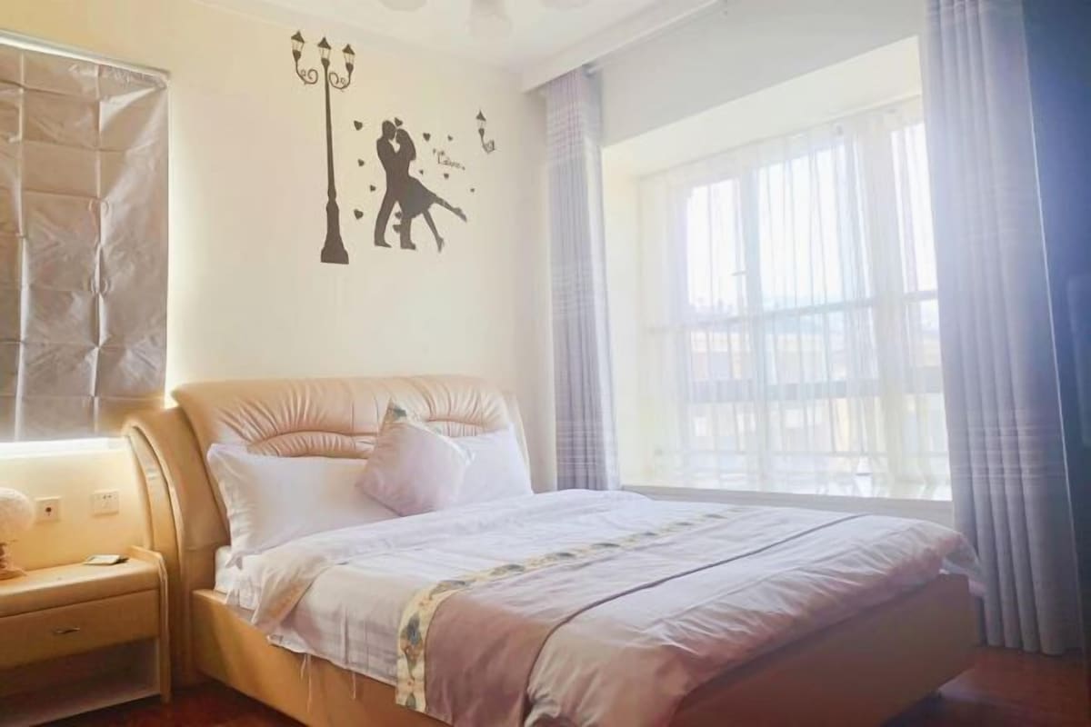 A bedroom with a bed, a nightstand, a wall decoration of a couple, and a window with curtains.