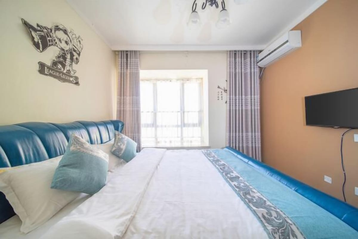 A bedroom with a blue sofa bed, a large bed with white bedding, a wall-mounted TV, and a window with curtains.