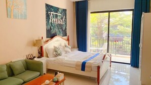 Un dormitorio con cama, sofá verde, mesita con bandeja, ventana con vista a follaje, y decoración de temática tropical.