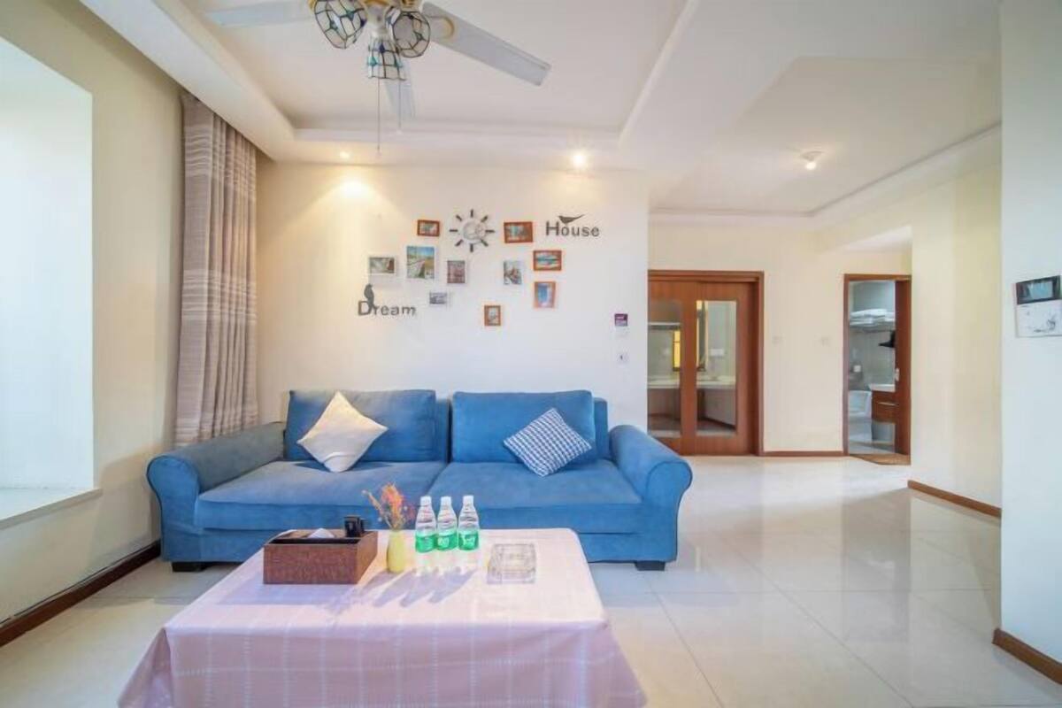 A living room with a blue sofa, a coffee table with drinks and a book, a ceiling fan, and a wall with framed pictures and words.