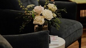 Un'area salotto con un vaso di fiori bianchi su un tavolino, un pianoforte sullo sfondo e un divano dai colori scuri.