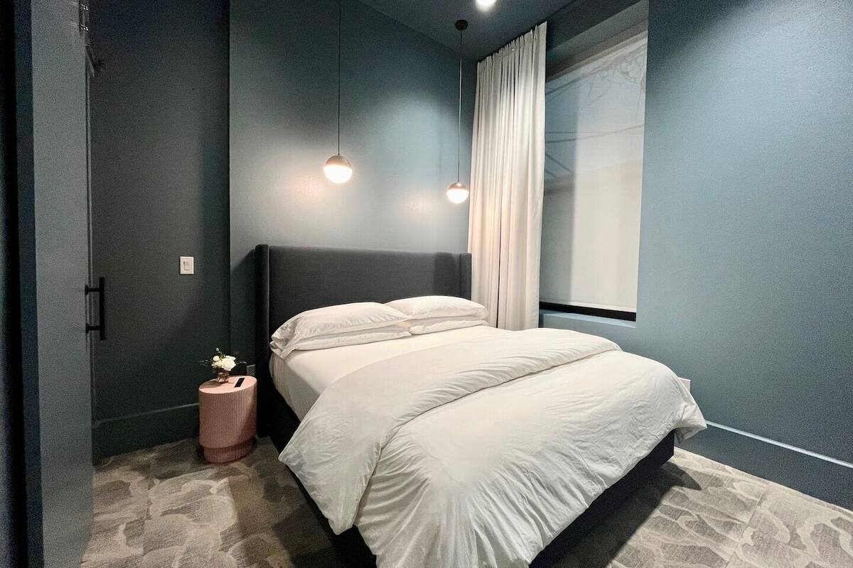 A bedroom with a bed, a nightstand, a small table with a plant, and a window with curtains.