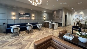 Un'accogliente hall d'hotel moderna con pavimento in legno a spina di pesce, una zona salotto centrale con divano e poltrone, un'area bar con bancone in marmo e un grande lampadario.