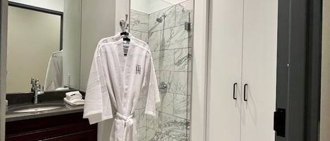 A modern bathroom with a marble floor, a glass shower enclosure, a white robe hanging on a rack, and a dark wooden vanity.