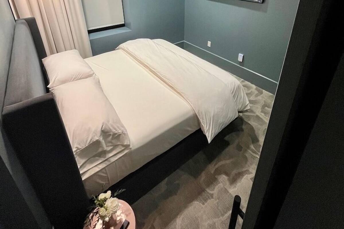 A bedroom with a bed, a bedside table with a lamp, a wall-mounted painting, and a window with curtains.