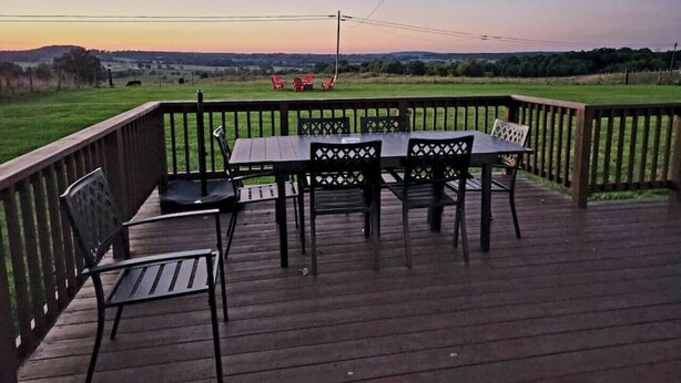 Una terraza de madera con una mesa y sillas, rodeada por una barandilla, con vistas a un paisaje cubierto de césped.