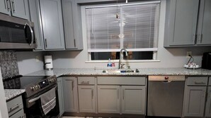 Una cocina con armarios grises, horno de acero inoxidable, lavavajillas y ventana con persianas.