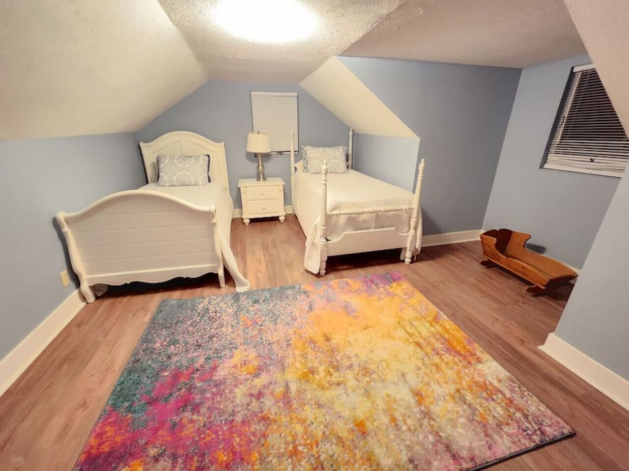 Ein Schlafzimmer mit schrägem Dach, zwei weißen Betten, einem farbenfrohen Teppich und einer Holzbank.
