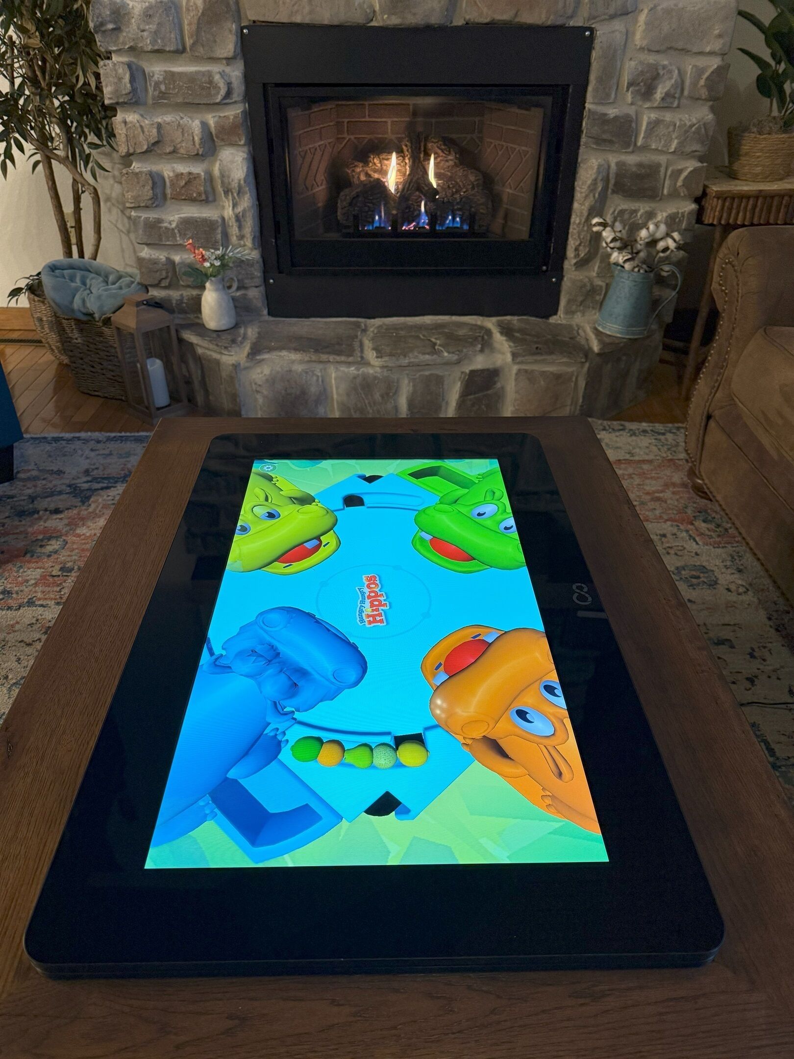 Une tablette affiche un jeu aux couleurs vives sur une table en bois dans un salon doté d’une cheminée en pierre.
