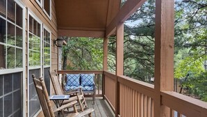 Un porche de madera con una zona de estar cubierta, una mesa y vistas a árboles.